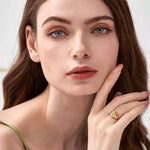 Portrait of a woman with blue eyes touching her cheek, wearing a gold embossed angel signet ring on her middle finger against a white background.