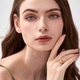 Portrait of a woman with blue eyes touching her cheek, wearing a gold embossed angel signet ring on her middle finger against a white background.