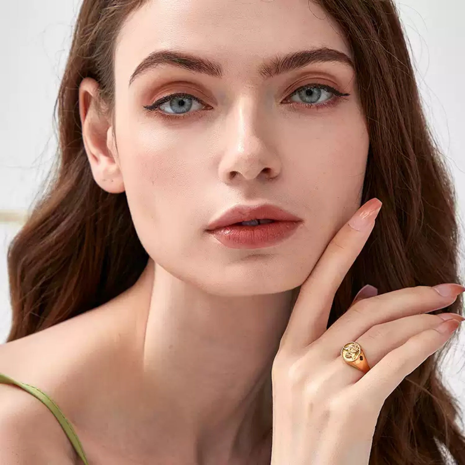 Portrait of a woman with blue eyes touching her cheek, wearing a gold embossed angel signet ring on her middle finger against a white background.