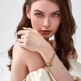 Portrait of a woman looking directly at the camera with her hand resting on her bare shoulder, wearing a gold asymmetric mixed chain bracelet with a dangling heart charm.