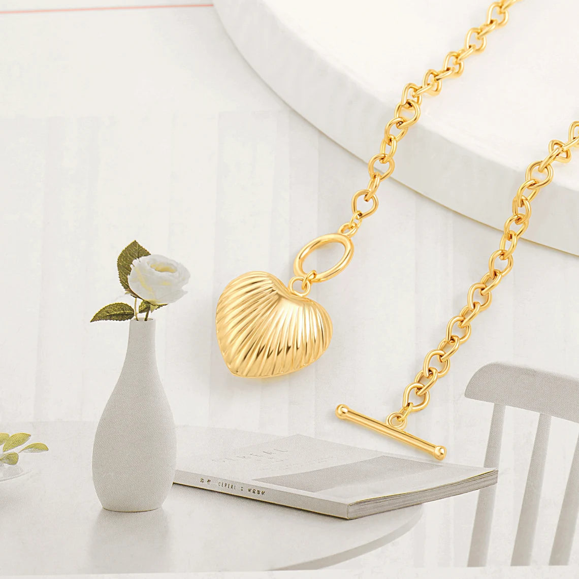 An aesthetic flat lay of a chunky gold toggle necklace with a ribbed heart pendant resting on a white table next to a small vase with a white rose.