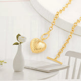 An aesthetic flat lay of a chunky gold toggle necklace with a ribbed heart pendant resting on a white table next to a small vase with a white rose.
