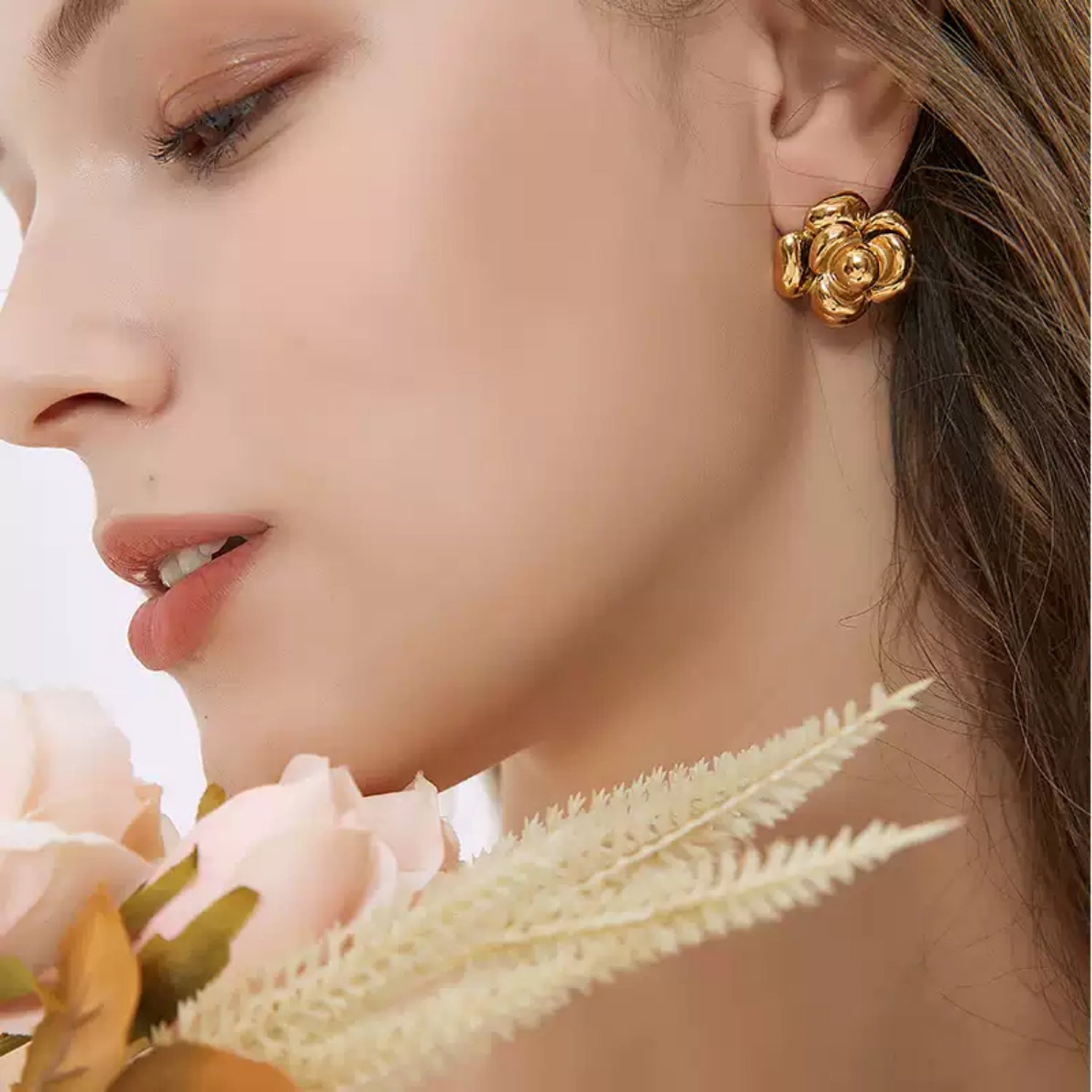 Side profile of a woman with her face close to pale pink faux flowers, displaying a shiny gold camellia flower stud earring.
