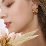 Side profile of a woman with her face close to pale pink faux flowers, displaying a shiny gold camellia flower stud earring.