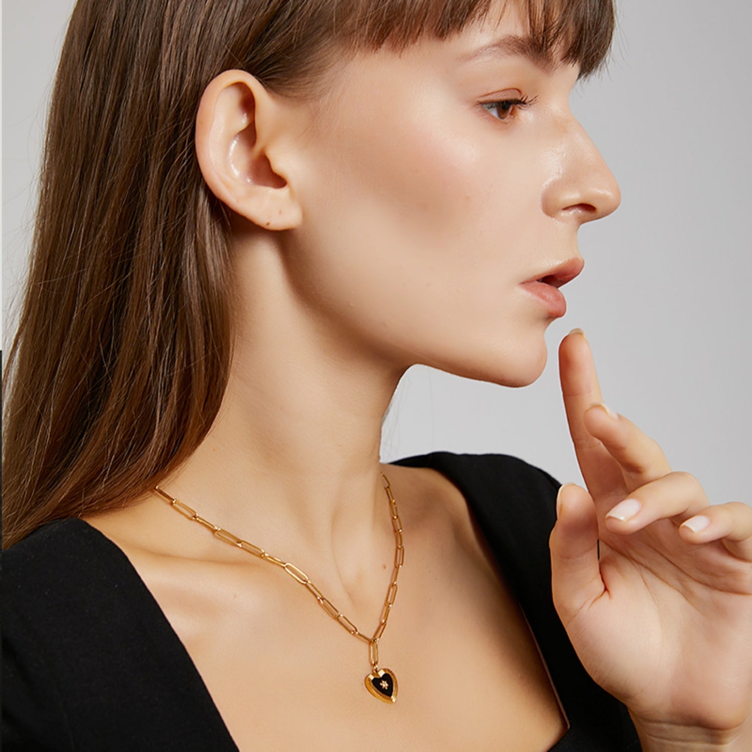 Side profile portrait of a woman wearing a black square-neck top, with her index finger touching her chin, showcasing a gold paperclip chain lariat necklace.