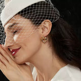 Side profile of a smiling woman with closed eyes wearing a white birdcage veil and a large gold hollow heart stud earring, with her hands clasped near her cheek.