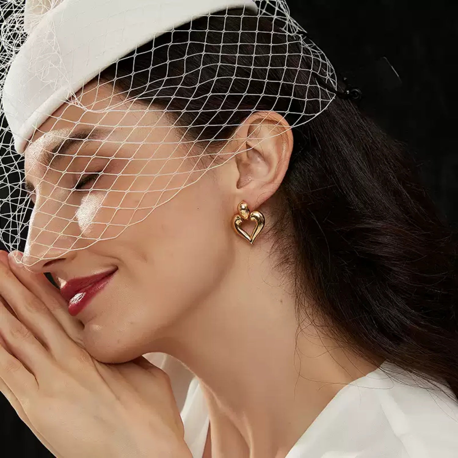 Side profile of a smiling woman with closed eyes wearing a white birdcage veil and a large gold hollow heart stud earring, with her hands clasped near her cheek.