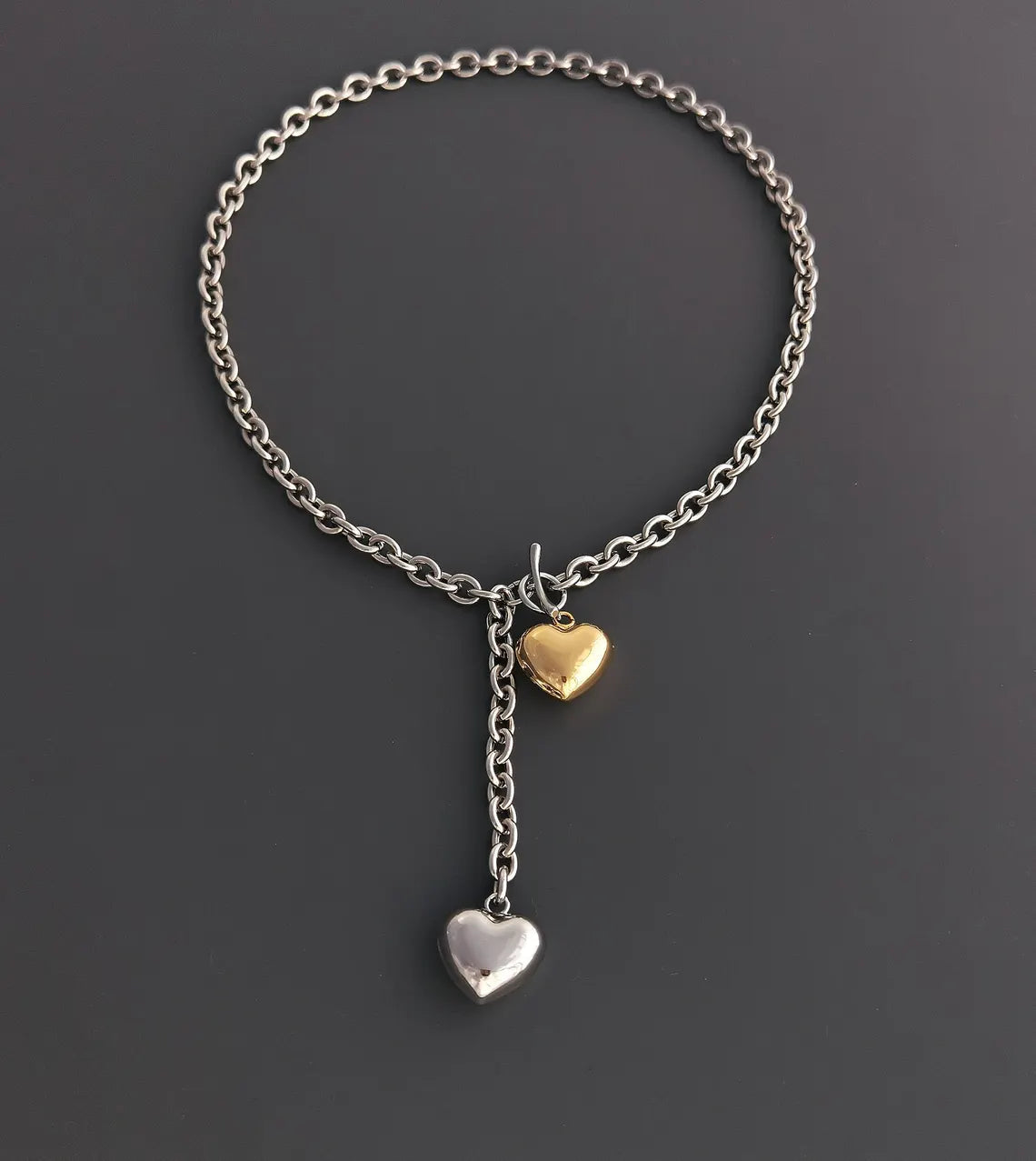 A flat lay view of a two-tone chunky cable chain necklace on a dark grey background, highlighting the front T-bar toggle clasp and puffy heart charms.