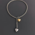 A flat lay view of a two-tone chunky cable chain necklace on a dark grey background, highlighting the front T-bar toggle clasp and puffy heart charms.