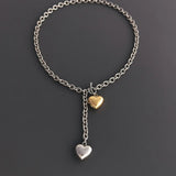 A flat lay view of a two-tone chunky cable chain necklace on a dark grey background, highlighting the front T-bar toggle clasp and puffy heart charms.