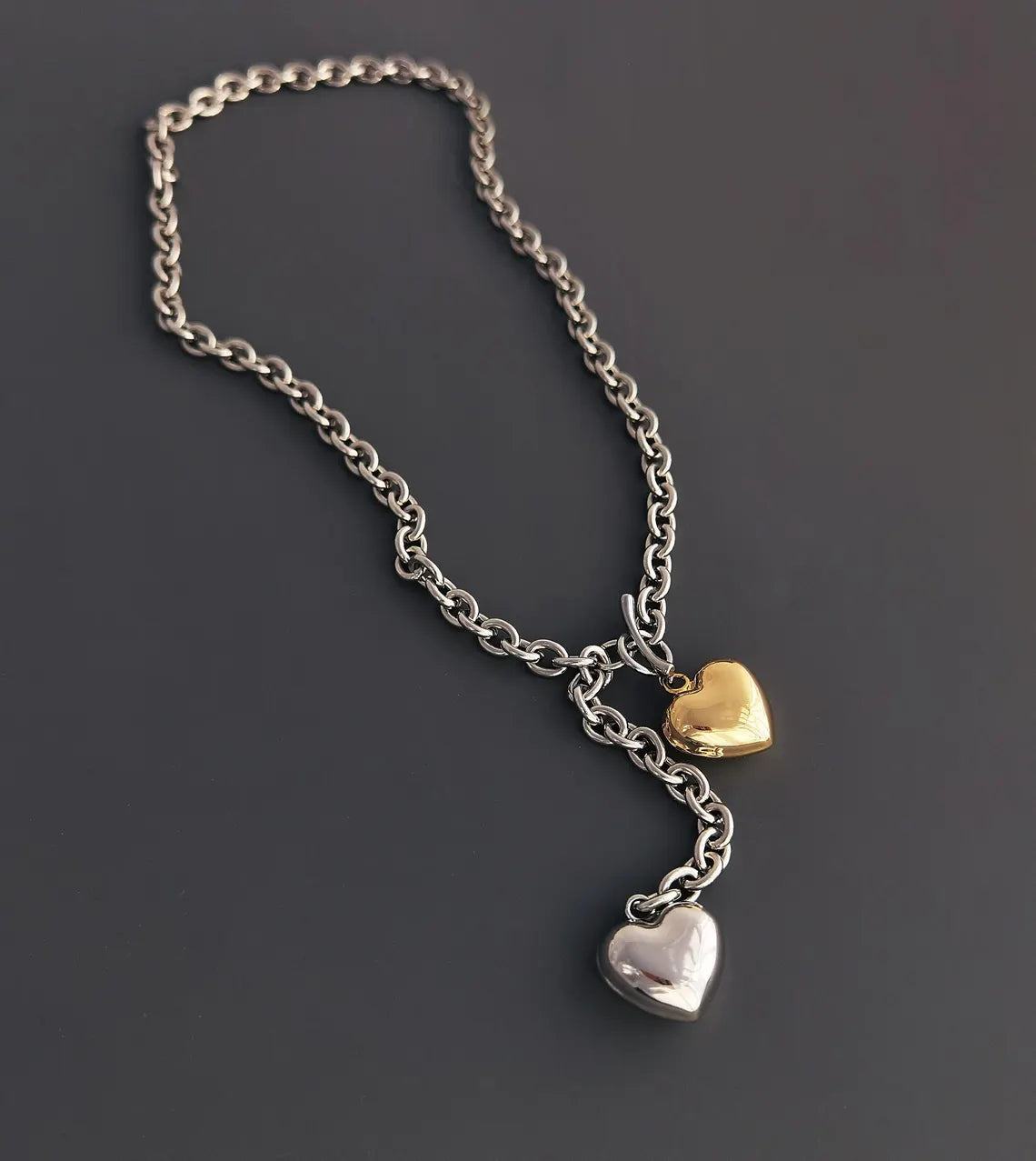 An angled flat lay shot of a mixed metal lariat necklace on a grey surface, showing the metallic shine of the silver and gold puffy heart pendants.