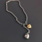 An angled flat lay shot of a mixed metal lariat necklace on a grey surface, showing the metallic shine of the silver and gold puffy heart pendants.