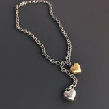 An angled flat lay shot of a mixed metal lariat necklace on a grey surface, showing the metallic shine of the silver and gold puffy heart pendants.
