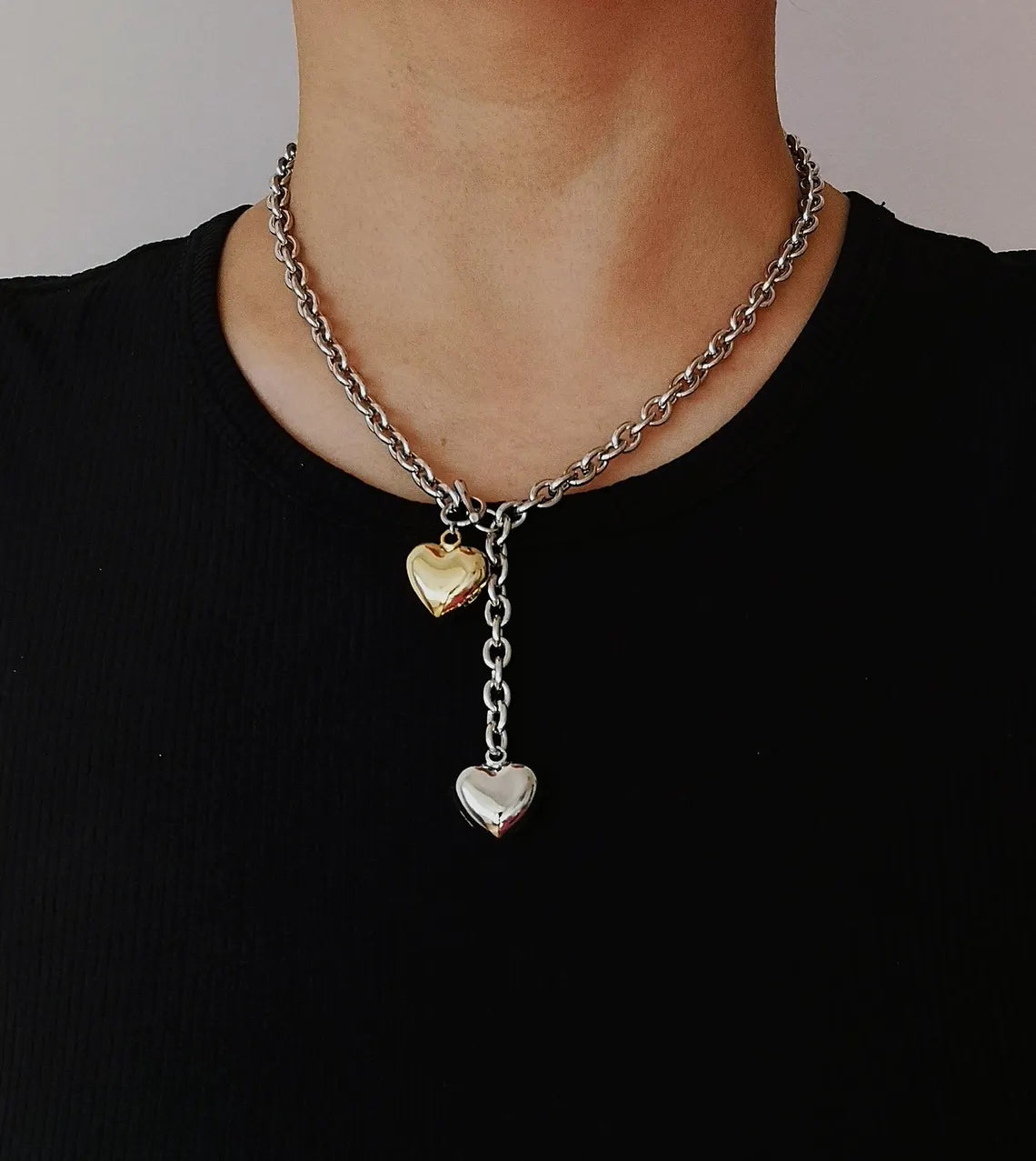 A close-up of a woman wearing a black top styled with a mixed metal chunky chain lariat necklace, featuring a front toggle and two 3D puffy heart pendants in silver and gold.