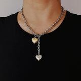 A close-up of a woman wearing a black top styled with a mixed metal chunky chain lariat necklace, featuring a front toggle and two 3D puffy heart pendants in silver and gold.