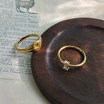 A dainty gold ring with a vertically set emerald-cut clear stone and a gold star signet ring, displayed on a dark round wooden coaster over a vintage printed book page.