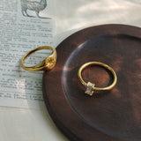 A dainty gold ring with a vertically set emerald-cut clear stone and a gold star signet ring, displayed on a dark round wooden coaster over a vintage printed book page.