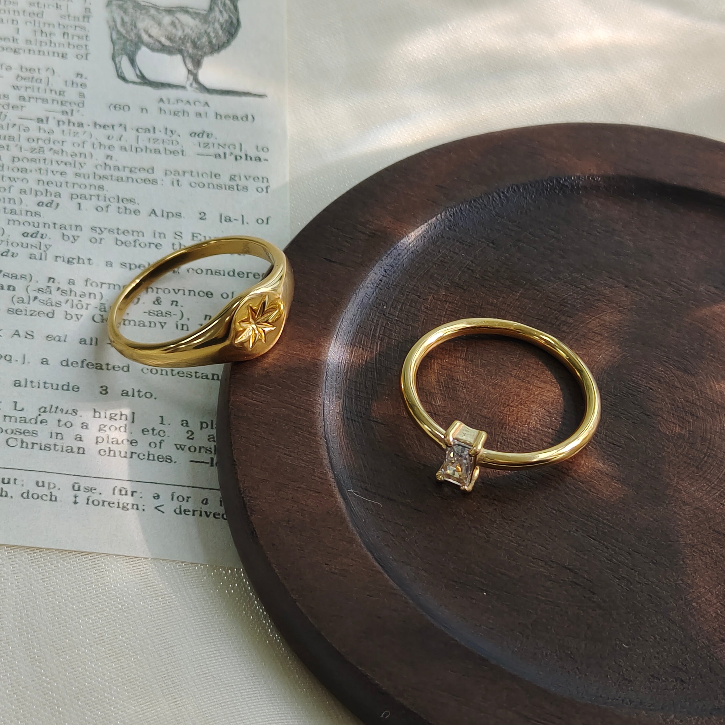 A dainty gold ring with a vertically set emerald-cut clear stone and a gold star signet ring, displayed on a dark round wooden coaster over a vintage printed book page.