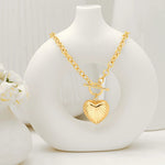 A vintage-inspired gold ribbed heart toggle necklace draped over a modern white circular vase with small white flowers in the background.