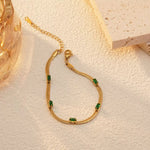 A gold plated snake chain bracelet with green rectangular stones laid on a textured white surface, next to a piece of porous stone and an amber glass object under warm sunlight.