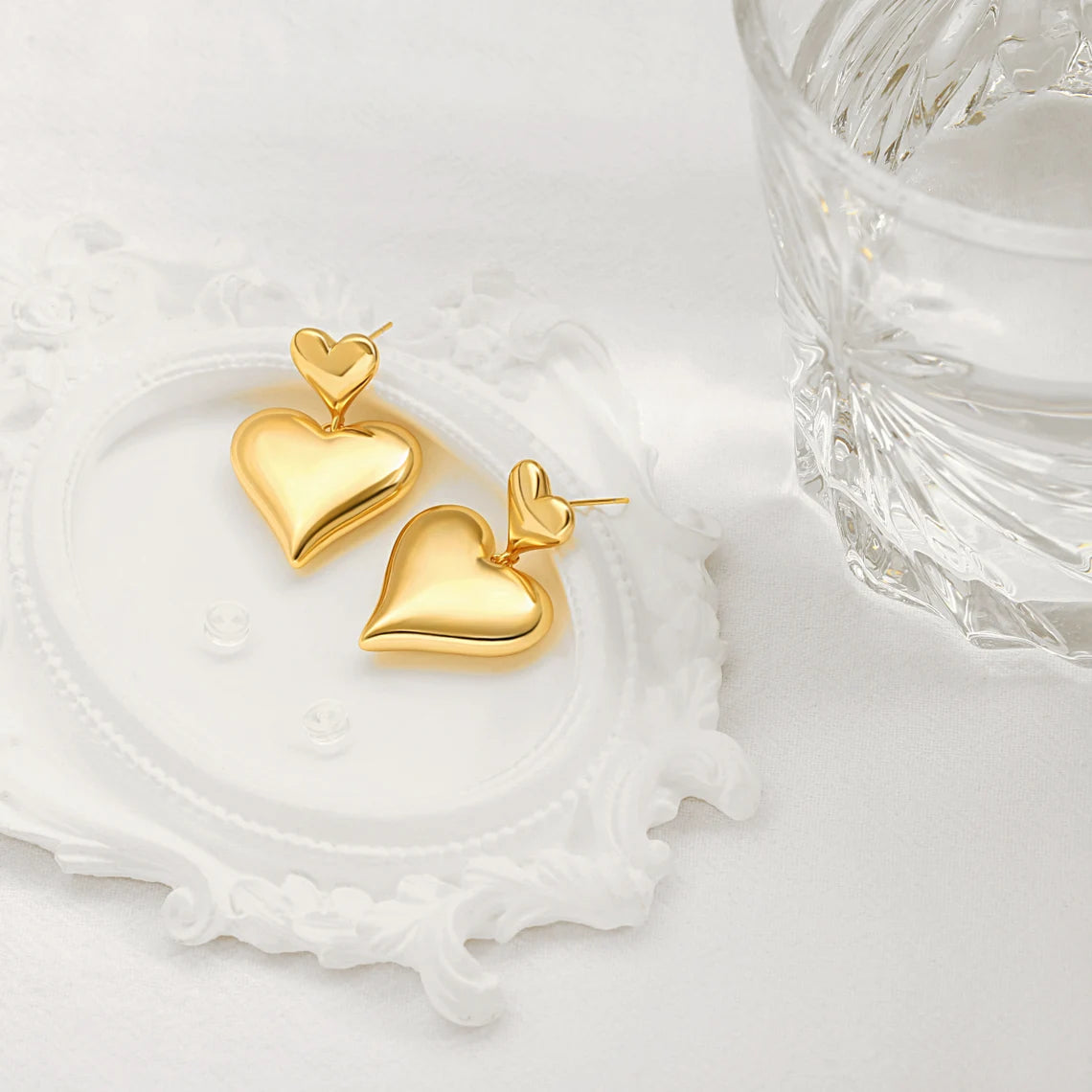 An aesthetic flat lay of two 18K gold puffy heart drop earrings resting on a white decorative vintage frame next to a textured glass.