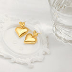 An aesthetic flat lay of two 18K gold puffy heart drop earrings resting on a white decorative vintage frame next to a textured glass.