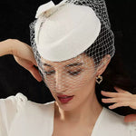 A woman wearing a white top and a white vintage fascinator hat with a birdcage veil looking downwards, showcasing a chunky gold openwork heart earring.