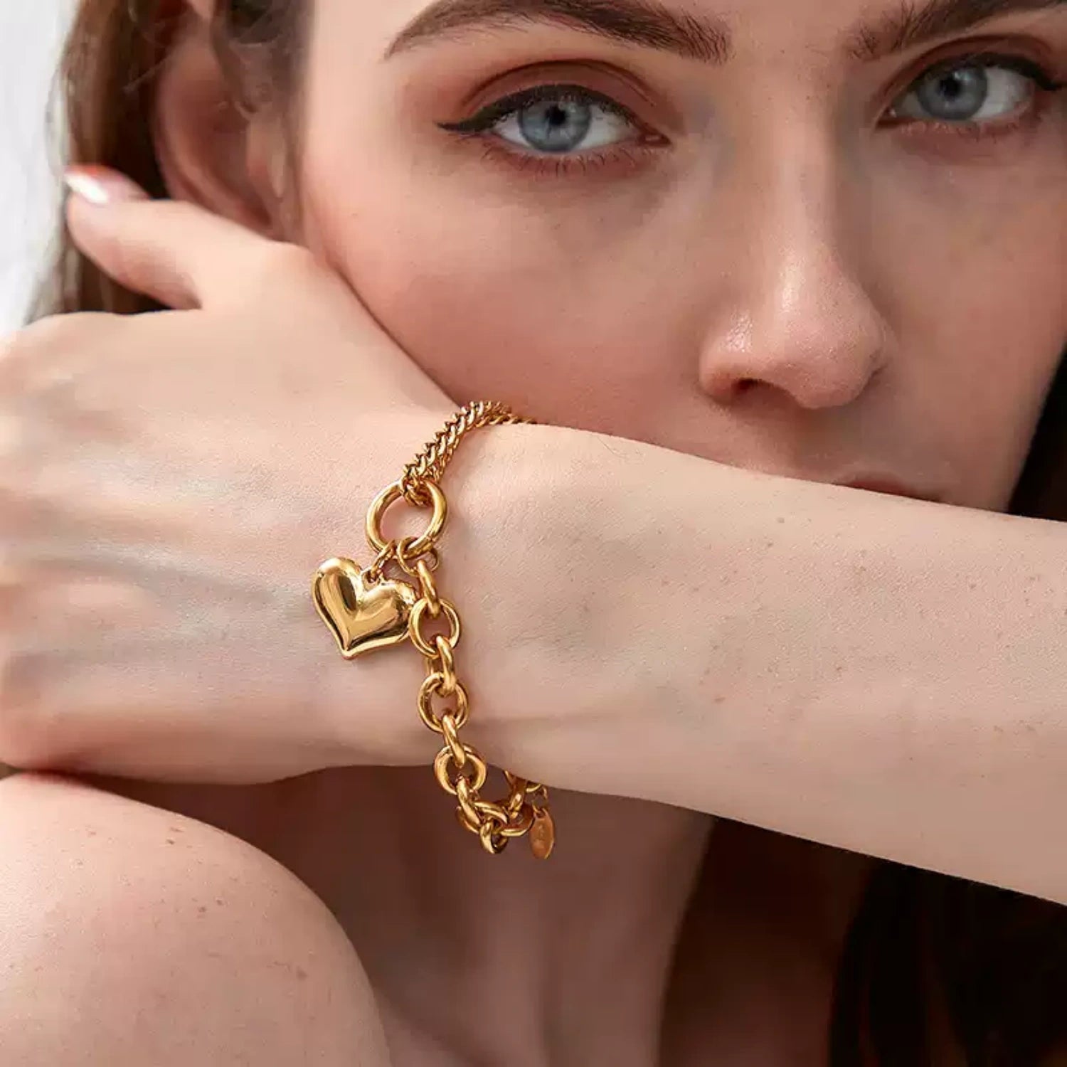 A woman with blue eyes resting her arm horizontally across her lower face, prominently displaying a shiny gold puffy heart charm bracelet on her wrist.