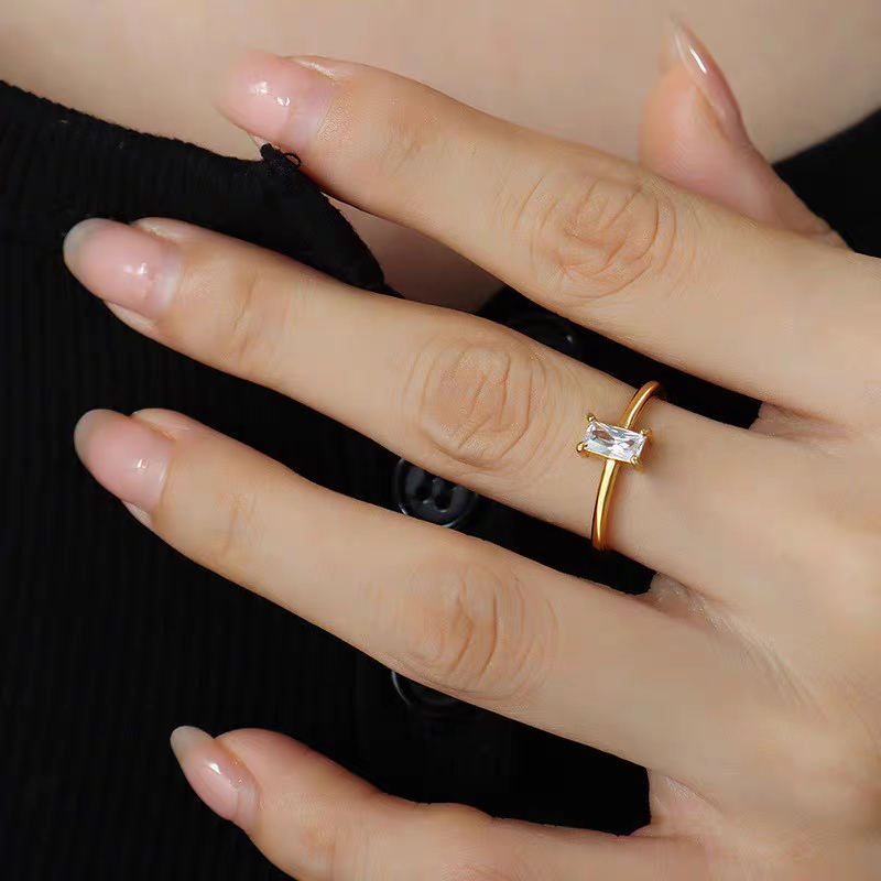Close-up of a woman's hand resting gracefully on a black ribbed garment, showcasing a dainty gold ring with a clear vertically set emerald-cut cubic zirconia.