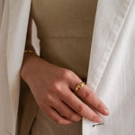 A woman wearing a beige top and a white blazer, elegantly showcasing a gold starburst signet ring on her ring finger.