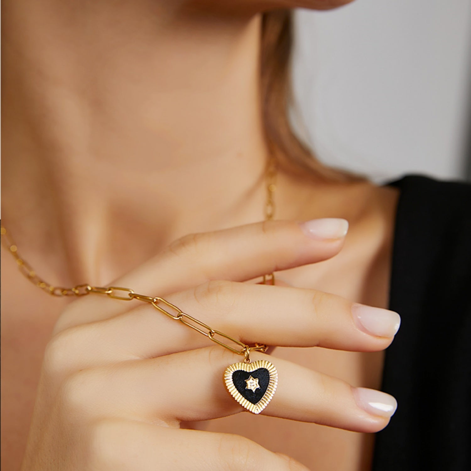 A woman wearing a black top with her hand gracefully resting on her collarbone, displaying an 18K gold plated Y-drop lariat necklace with a black enamel heart.