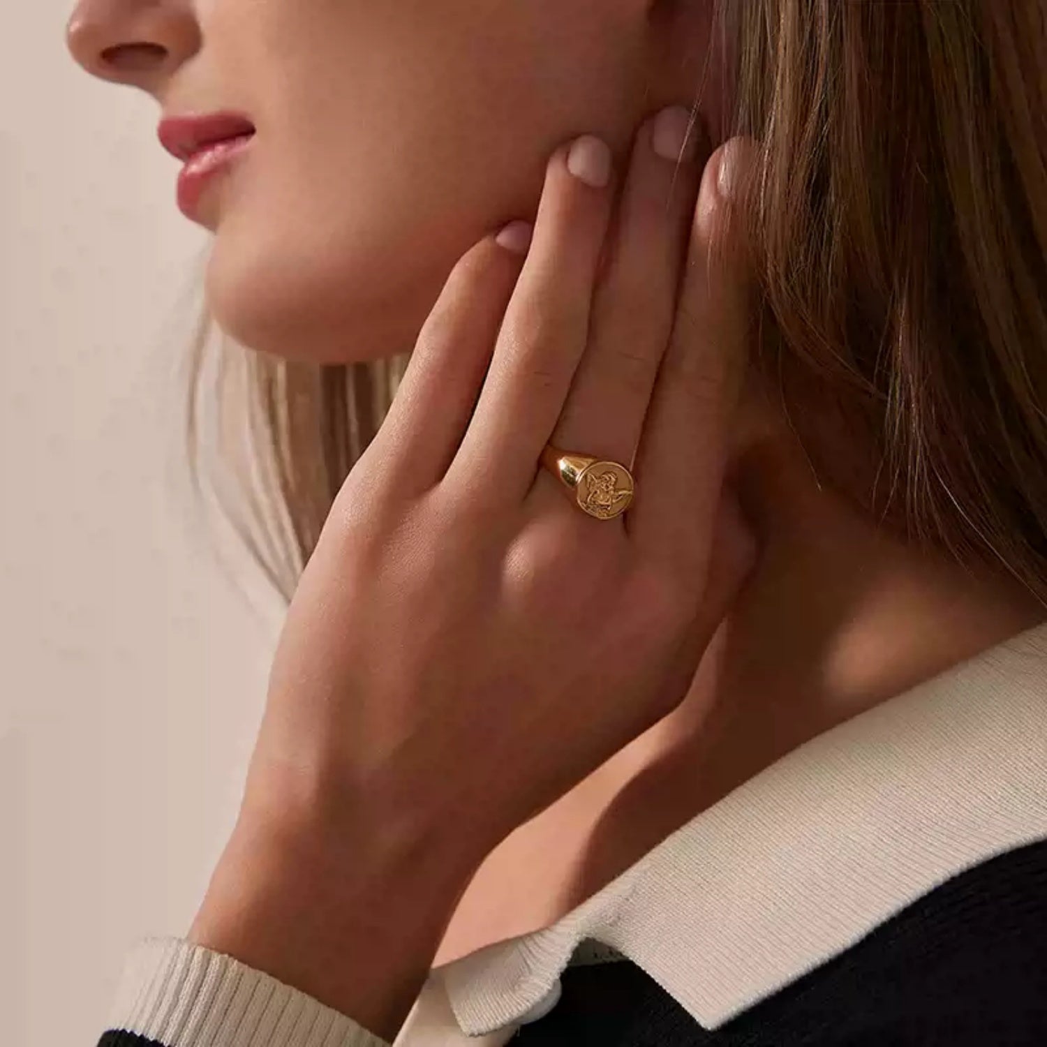 Close-up profile of a woman touching her neck, wearing a gold signet ring with an embossed angel design on her hand.