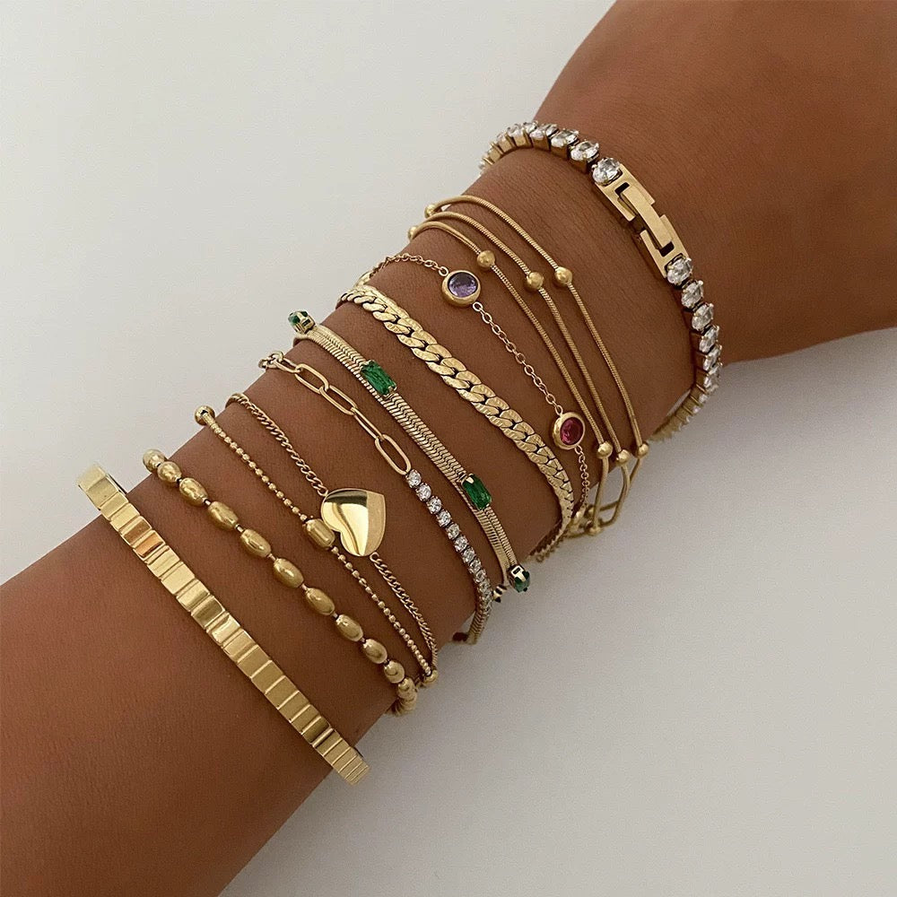 A woman's arm with a deep tan modeling a large curated stack of various gold bracelets, including a tennis bracelet, bead bracelets, cuban links, and the gold emerald baguette station bracelet in the center.