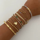 A woman's arm with a deep tan modeling a large curated stack of various gold bracelets, including a tennis bracelet, bead bracelets, cuban links, and the gold emerald baguette station bracelet in the center.